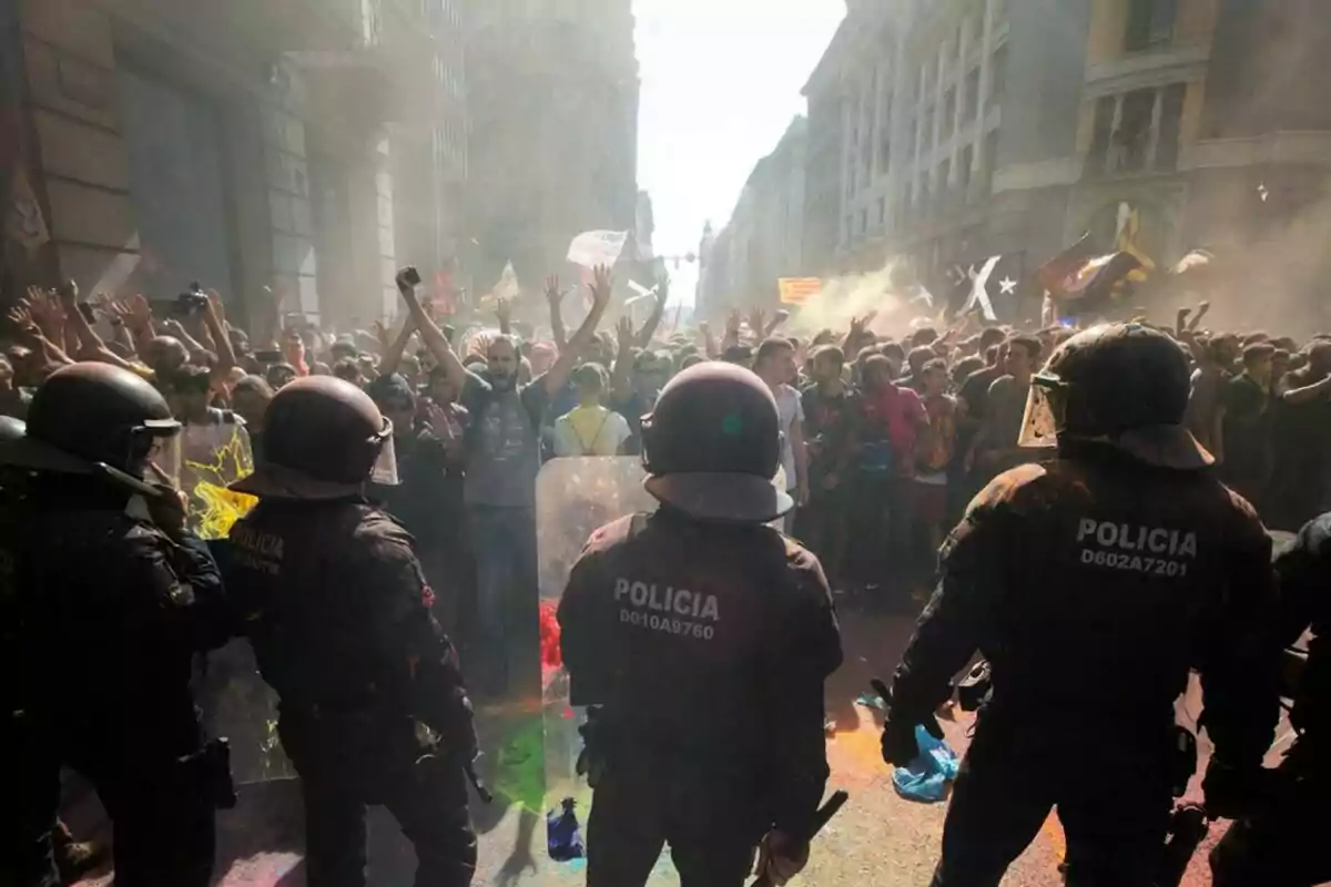 Una multitud de manifestants s'enfronta a un grup de policies antiavalots en un carrer urbà, amb fum i colors a l'aire.