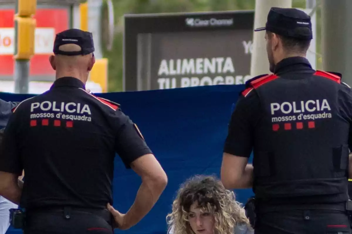 Dos agents de la policia dels Mossos d'Esquadra estan d'esquena, mentre una dona amb cabells arrissats es troba al centre de la imatge. Dos agents de la policia dels Mossos d'Esquadra estan d'esquena, mentre una dona amb cabells arrissats es troba al centre de la imatge.