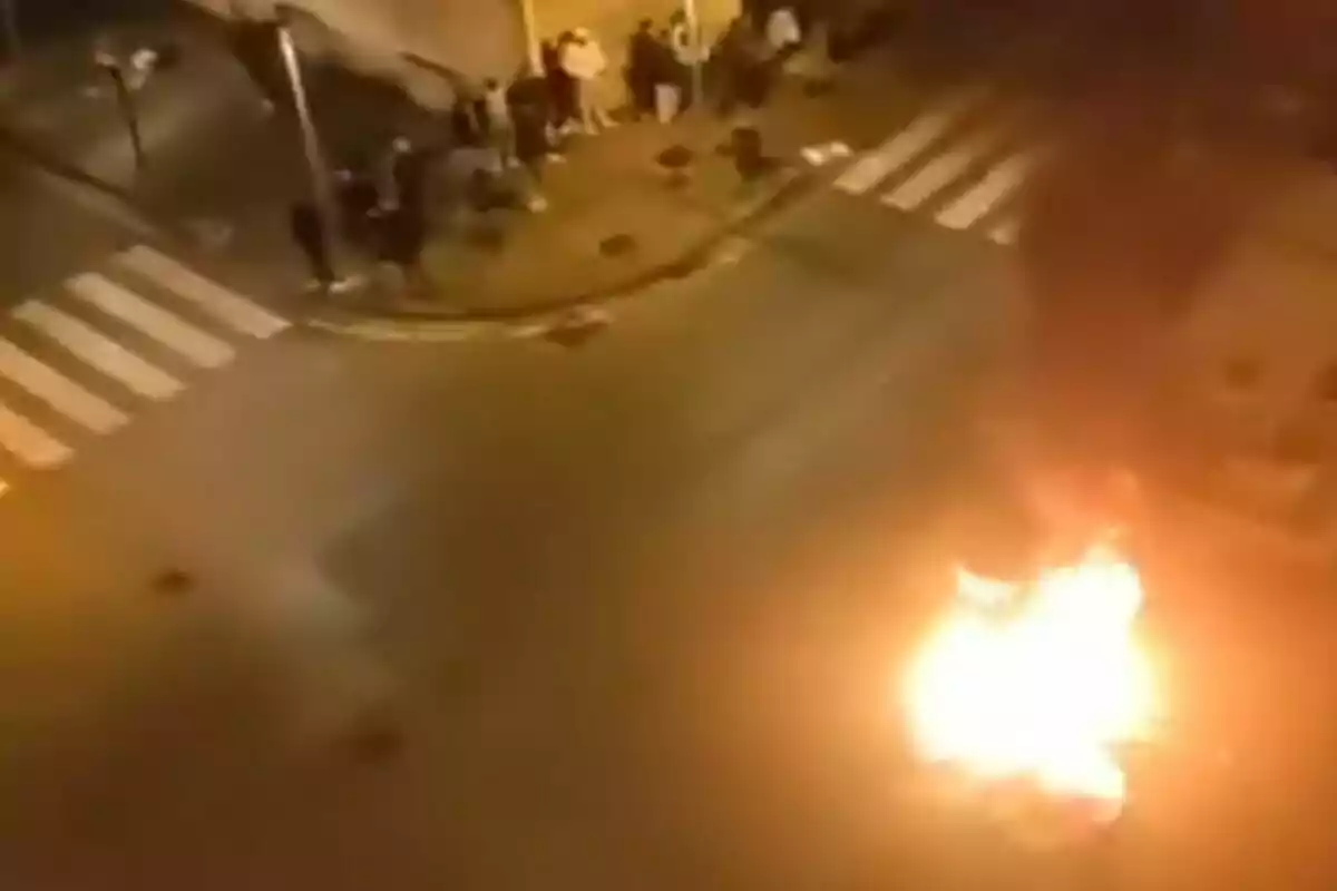 Una esquina de una calle por la noche con un grupo de personas observando un fuego en el pavimento.