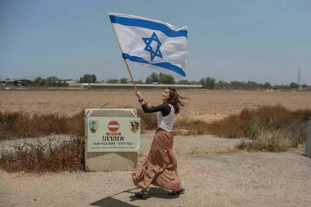Dona somrient onejant una bandera d’Israel al costat d’un cartell d’advertència en un camí rural Dona somrient onejant una bandera d’Israel al costat d’un cartell d’advertència en un camí rural
