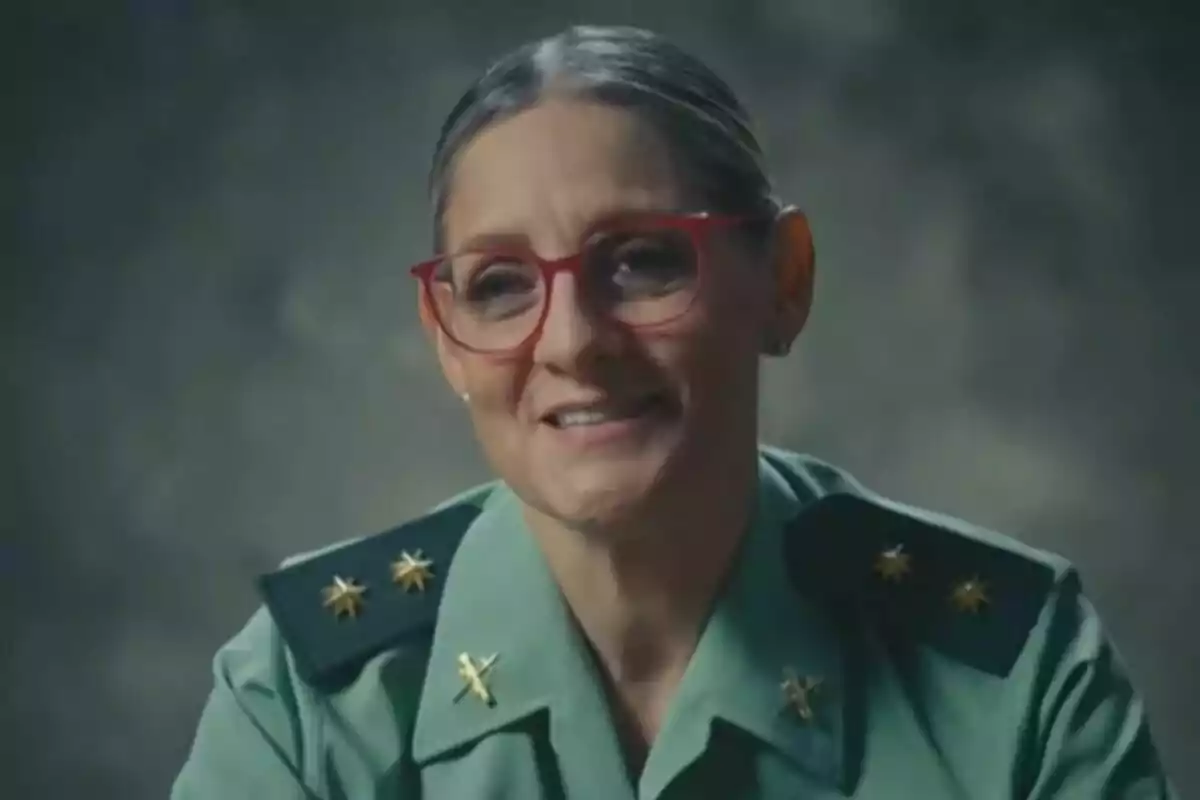 Una persona con gafas rojas y uniforme verde sonríe frente a un fondo oscuro en la promo de En Guardia: Mujeres contra el crimen.