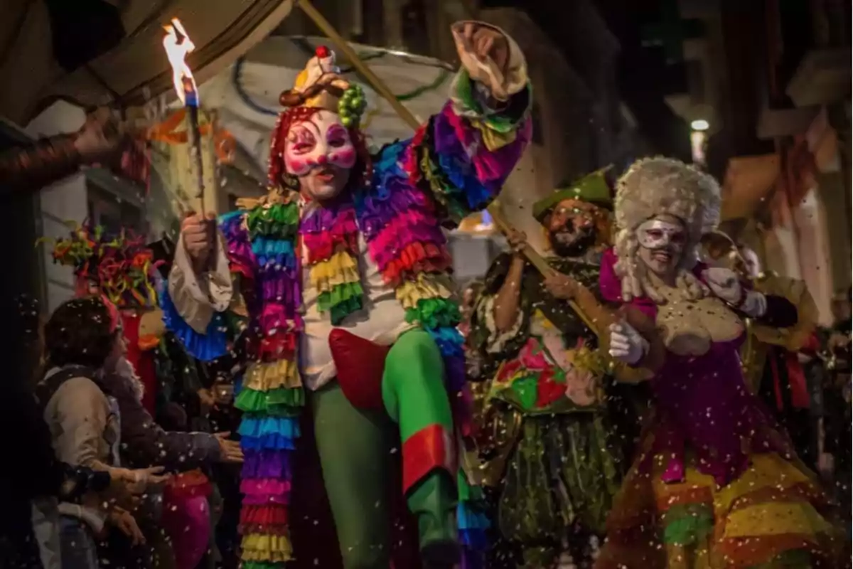 Personas disfrazadas con trajes coloridos y máscaras participan en un desfile festivo nocturno, rodeadas de confeti y luces.