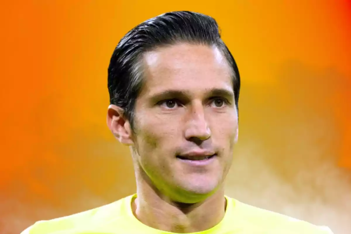 A man with dark, slicked-back hair, wearing a yellow t-jersey, against a gradient background in shades of orange.