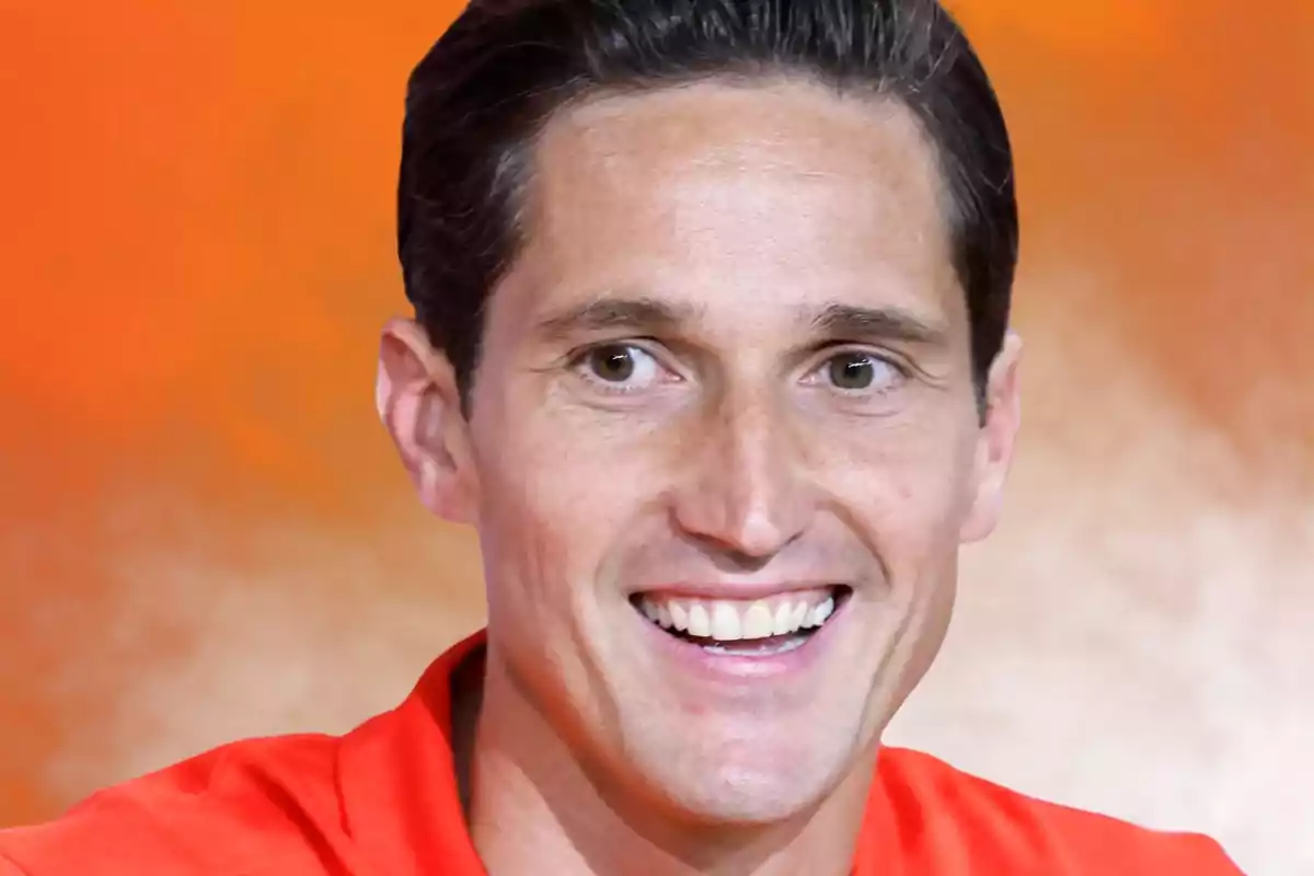 A man smiling with a red jersey and an orange background.