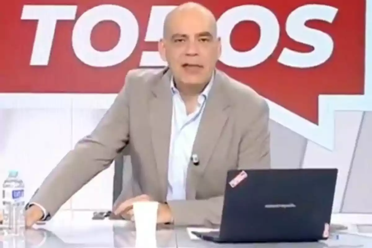 Nacho Abad amb vestit clar està assegut en un escriptori amb un portàtil i una ampolla d'aigua, davant d'un fons vermell amb lletres blanques a 'En boca de todos'.
