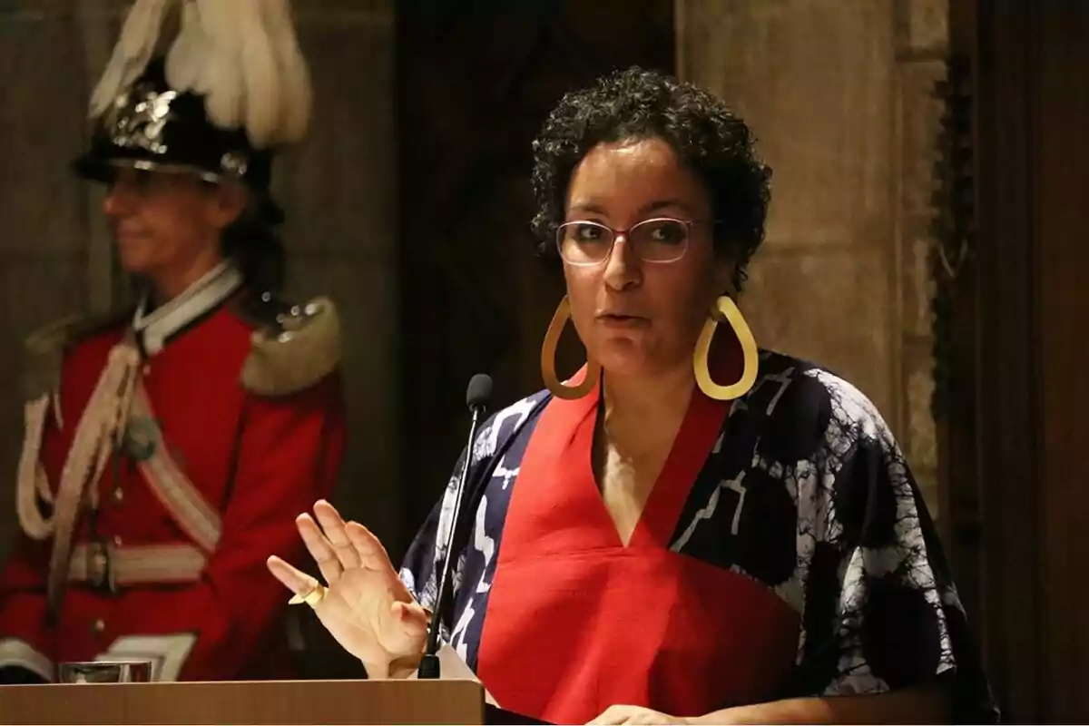 Una persona amb ulleres i arracades grans parla en un micròfon, mentre un guàrdia amb uniforme vermell i barret amb plomes està dret al fons. Una persona amb ulleres i arracades grans parla en un micròfon, mentre un guàrdia amb uniforme vermell i barret amb plomes està dret al fons.
