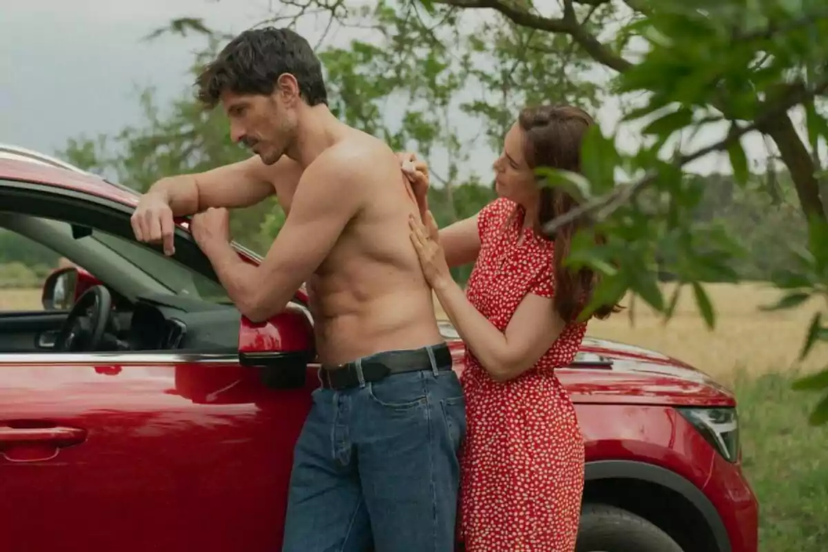 Andrés Velencoso sense camisa es recolza en un cotxe vermell mentre una Natalia Sánchez en vestit vermell li aplica alguna cosa a l'esquena en un entorn exterior a Retorn a les Sabines.