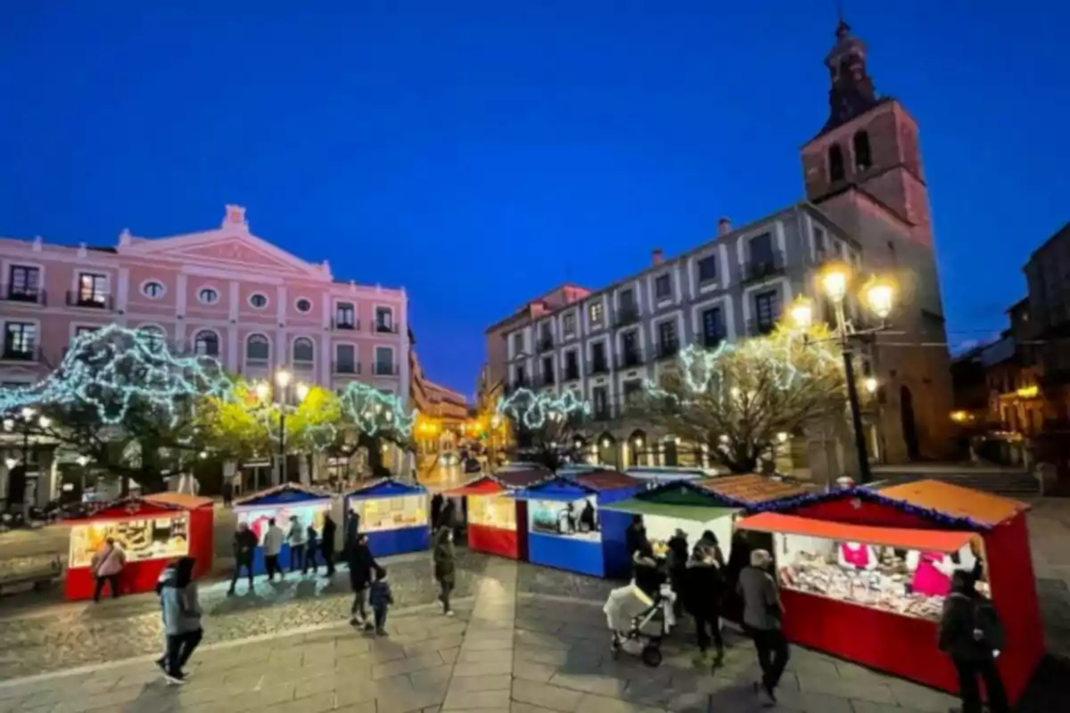 Mercat nadalenc a l'aire lliure amb casetes il·luminades i persones passejant a una plaça amb edificis històrics i arbres decorats amb llums.