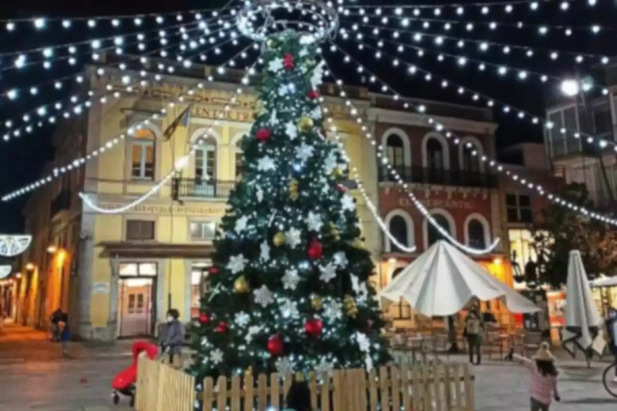 Arbre de Nadal il·luminat en una plaça amb llums penjants i edificis de fons.