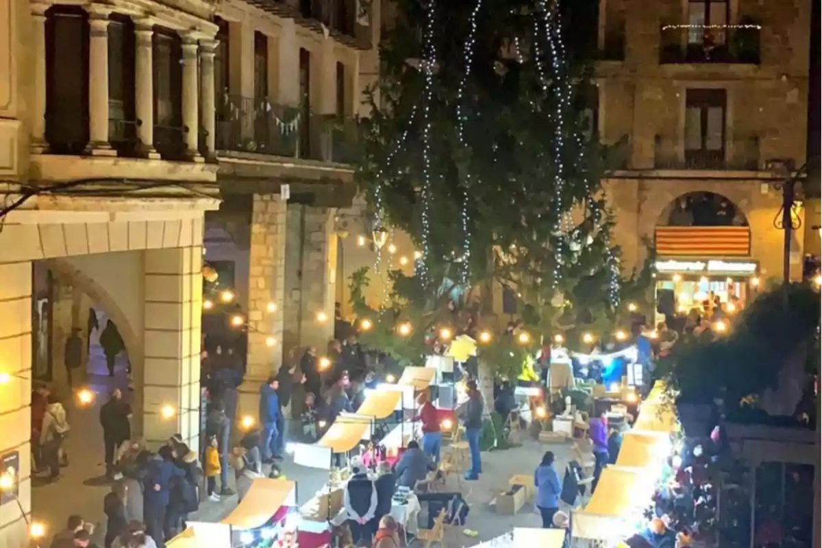 Mercat nadalenc nocturn amb llums brillants i parades decorades en una plaça envoltada d'edificis històrics.