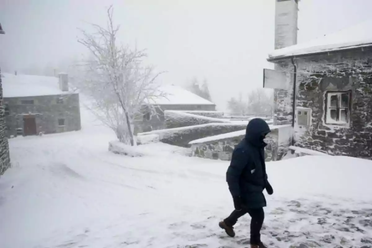 Una persona camina per un poble cobert de neu amb cases de pedra i un arbre sense fulles.