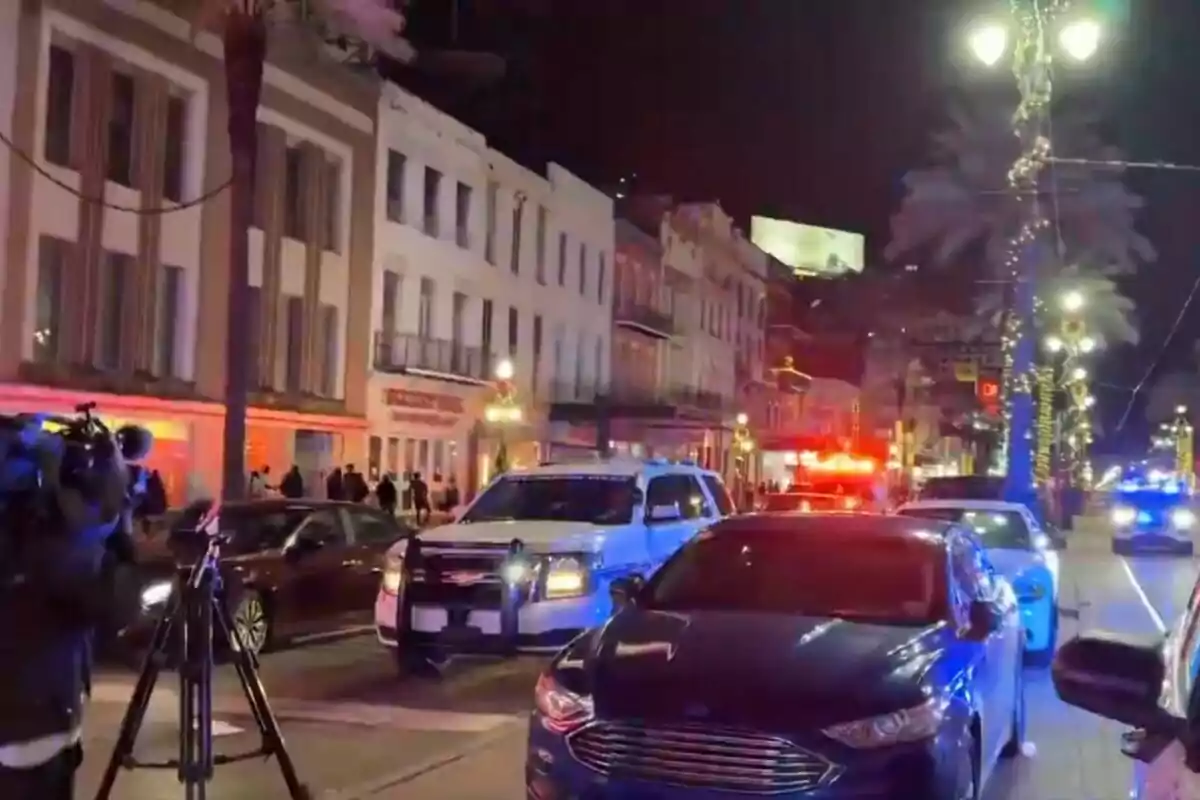 Una calle iluminada por la noche con vehículos de policía y cámaras de televisión presentes.