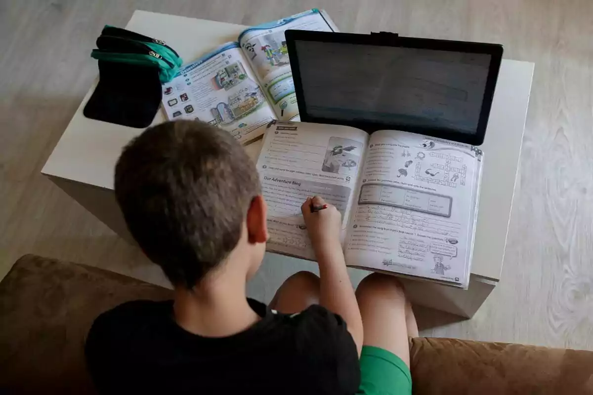 Un nen està assegut en un sofà fent deures escolars amb un llibre obert sobre les cames, una tauleta davant seu i més llibres i estoigs sobre una taula blanca. Un nen està assegut en un sofà fent deures escolars amb un llibre obert sobre les cames, una tauleta davant seu i més llibres i estoigs sobre una taula blanca.