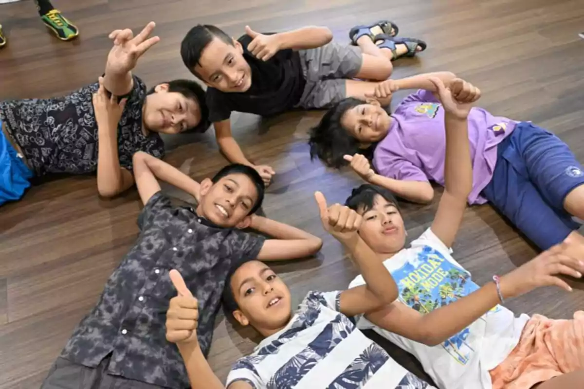 Un grupo de niños acostados en el suelo de madera, sonriendo y levantando los pulgares.