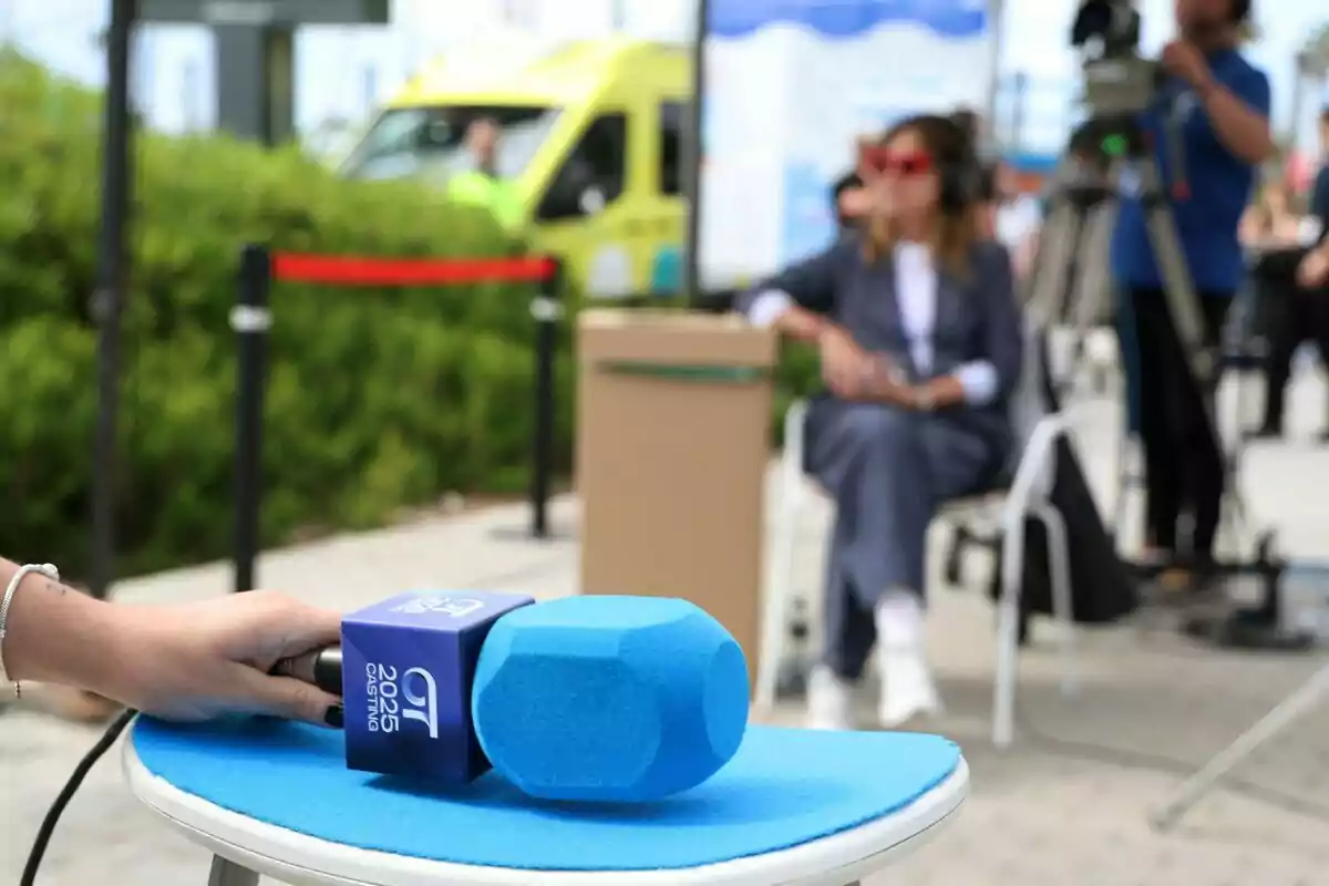Una mà sosté un micròfon blau amb un logotip mentre al fons es veu la Noemí Galera asseguda i una càmera de vídeo en un entorn a l'aire lliure en el càsting d'OT 2025. Una mà sosté un micròfon blau amb un logotip mentre al fons es veu la Noemí Galera asseguda i una càmera de vídeo en un entorn a l'aire lliure en el càsting d'OT 2025.