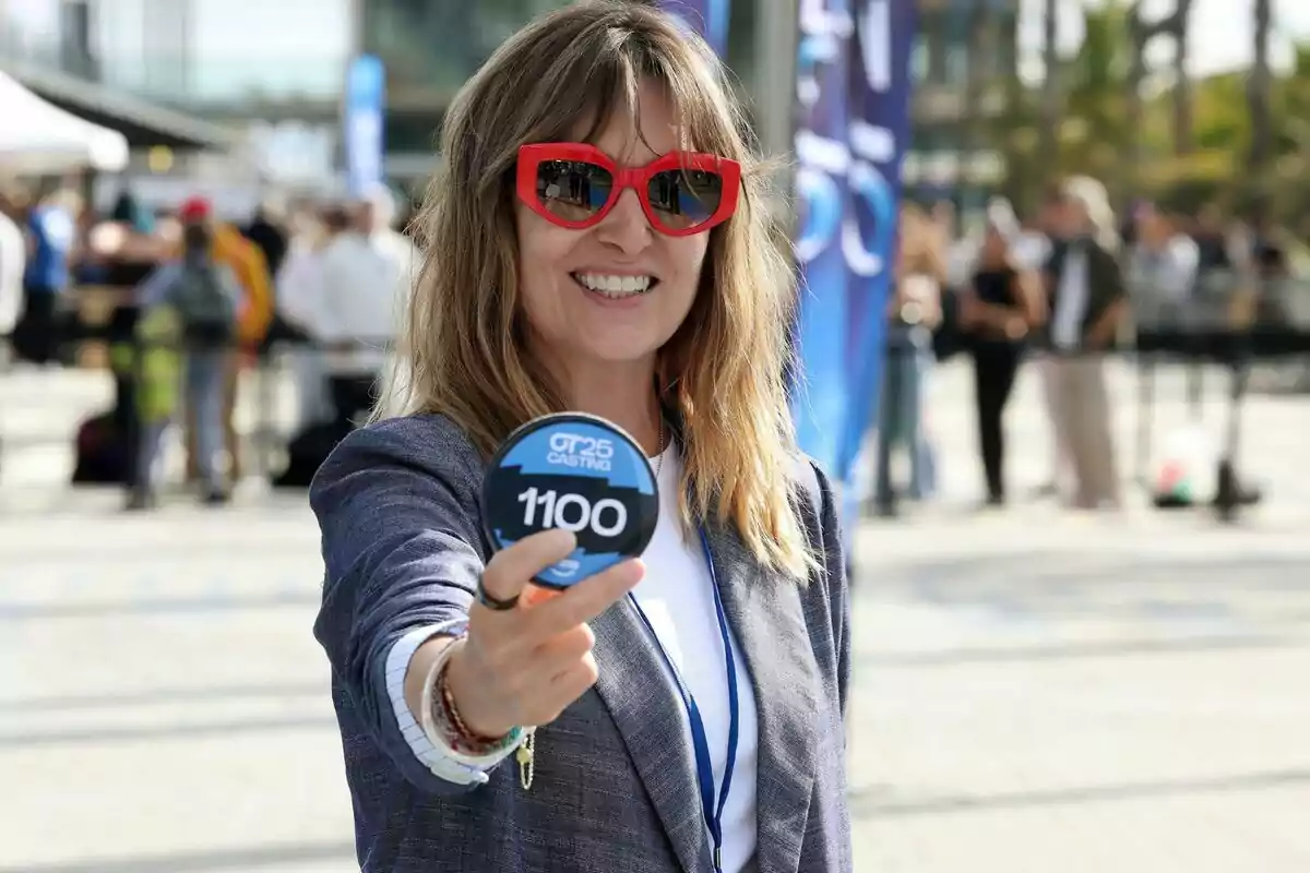 Noemí Galera somrient amb ulleres de sol vermelles sosté un cartell rodó amb el número 1100 en un esdeveniment a l'aire lliure en el càsting d'OT 2025.