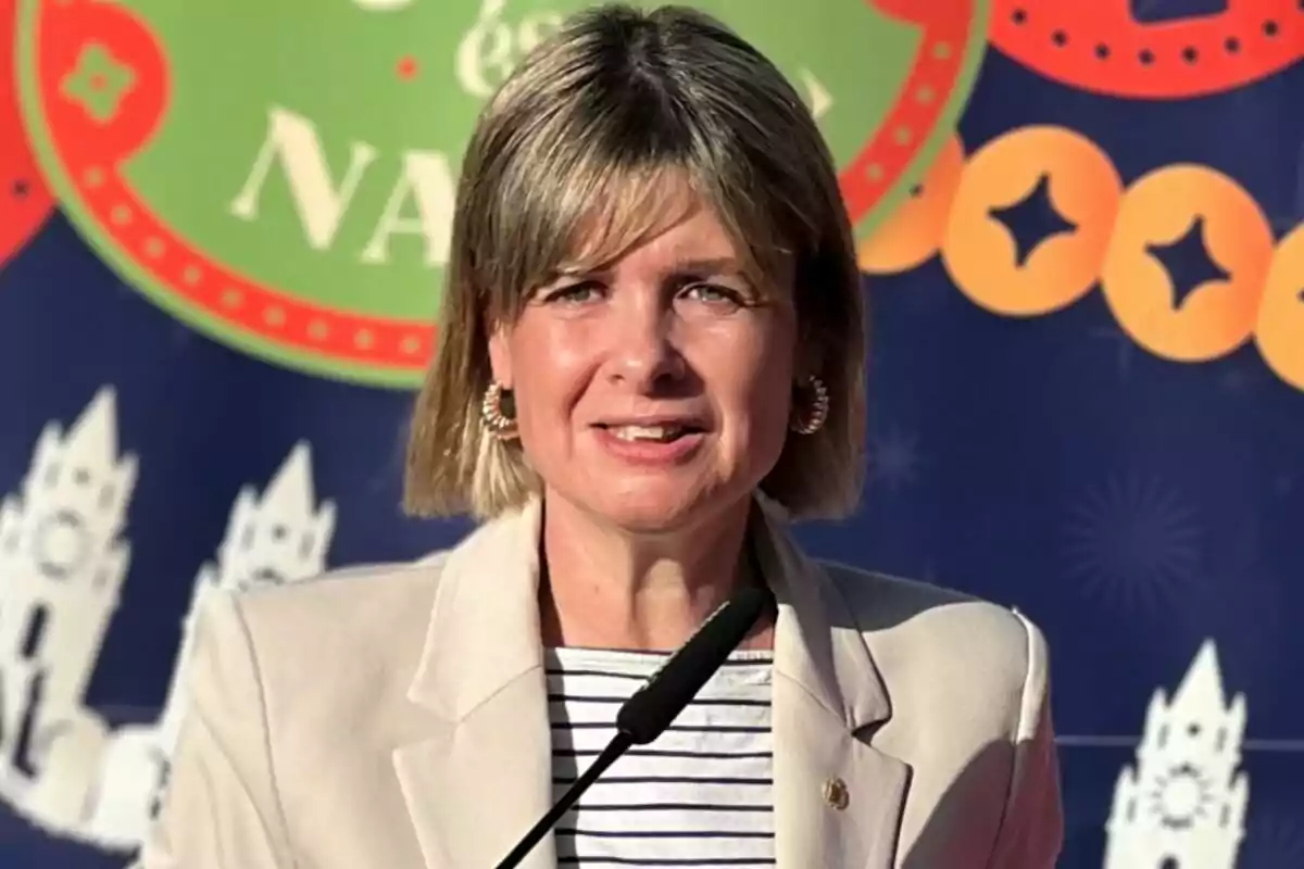 Noemí Llauradó with blonde hair and a light jacket speaking in front of a microphone with a colorful background.