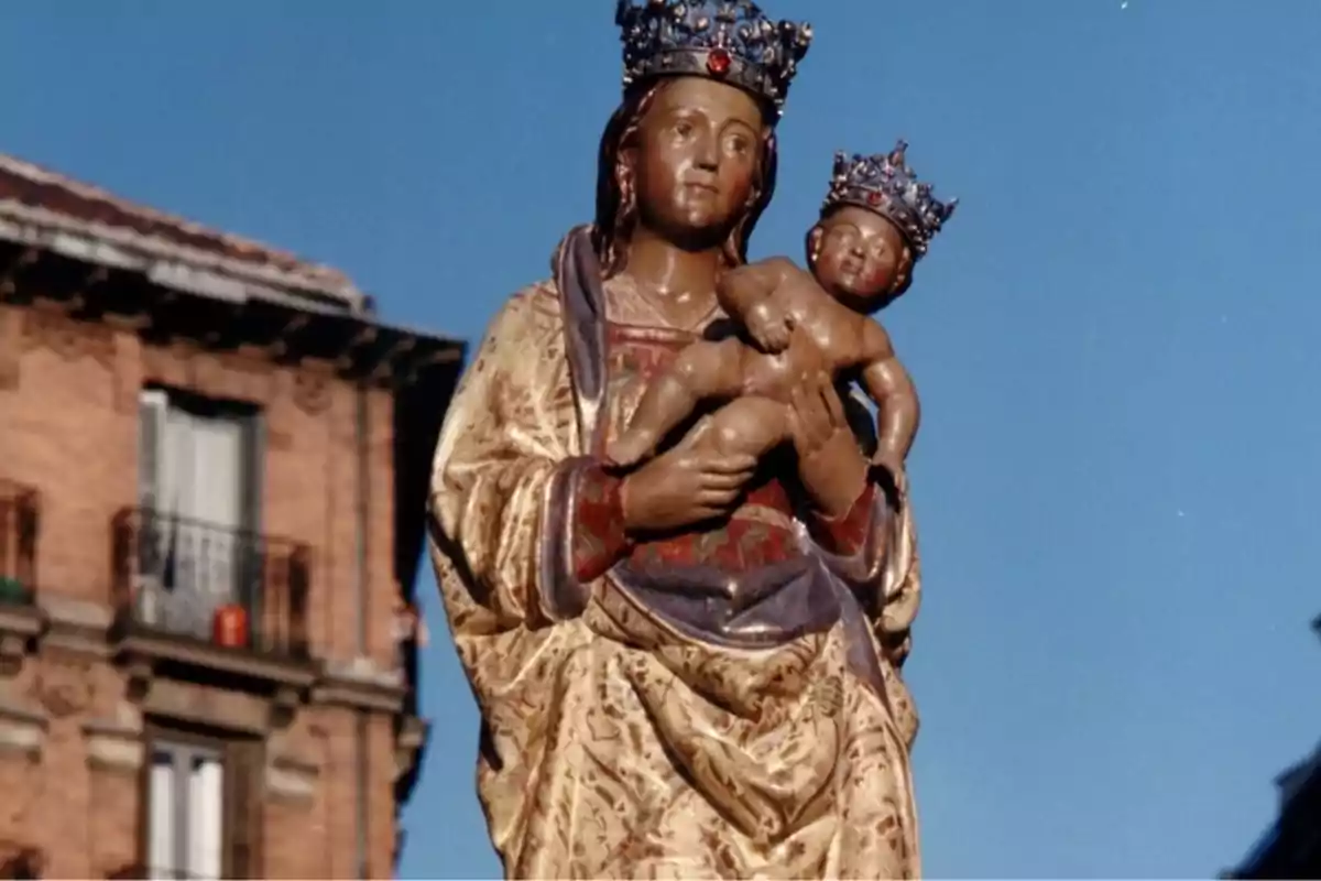 Escultura d'una figura religiosa amb un nen als braços i corones als dos caps, amb un edifici de maó al fons.