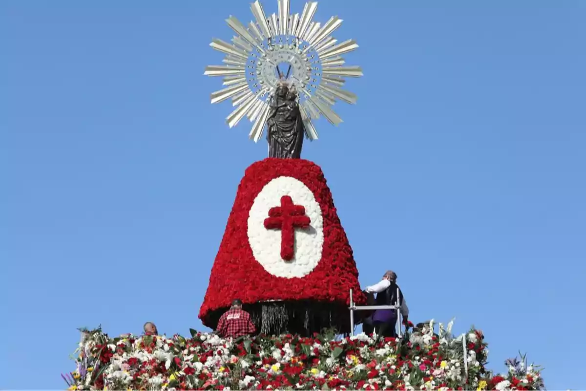 Una estàtua religiosa sobre una estructura coberta de flors vermelles i blanques amb una creu vermella al centre i un cel blau de fons.