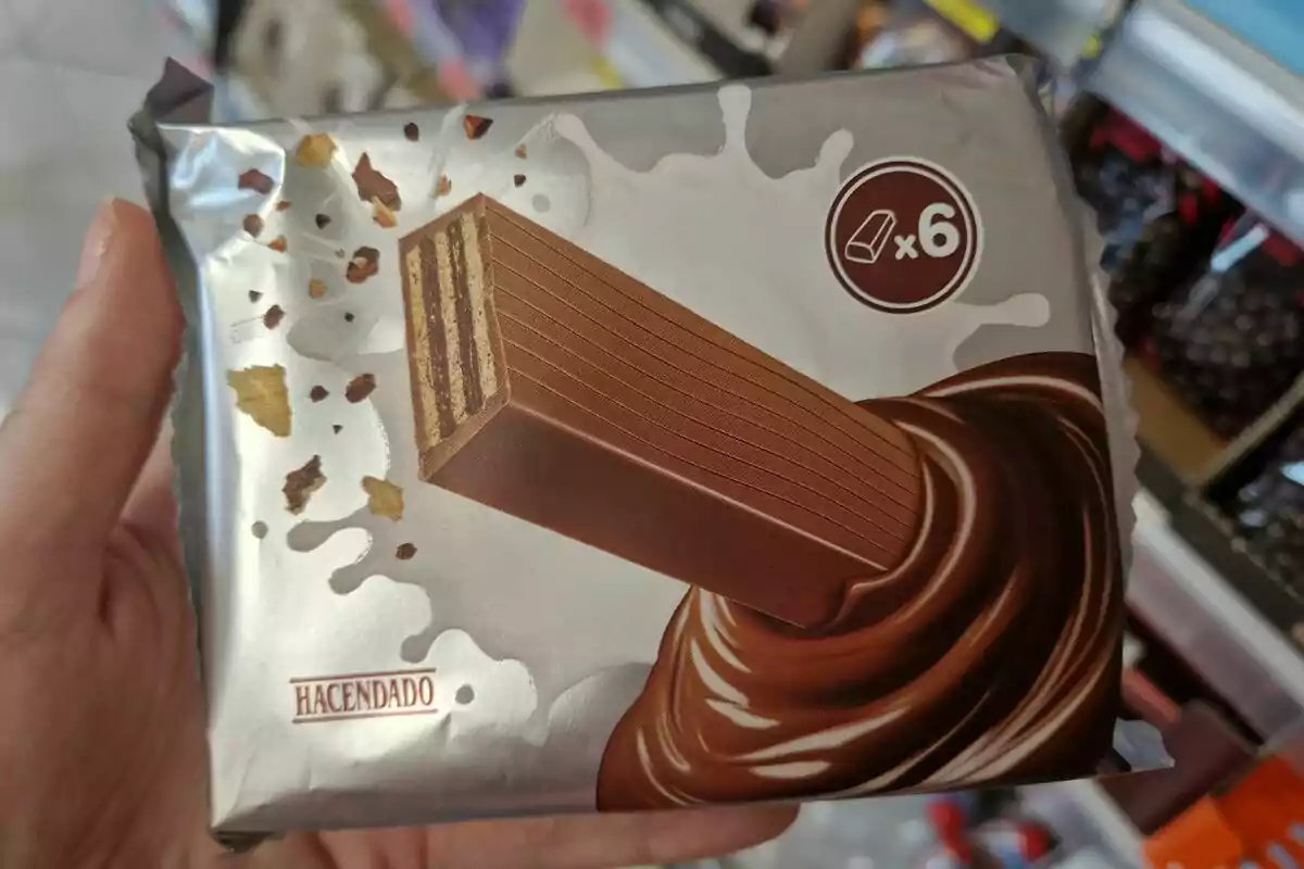 Una mano sostiene un paquete de barritas de chocolate de la marca Hacendado, que contiene seis unidades. Una mano sostiene un paquete de barritas de chocolate de la marca Hacendado, que contiene seis unidades.