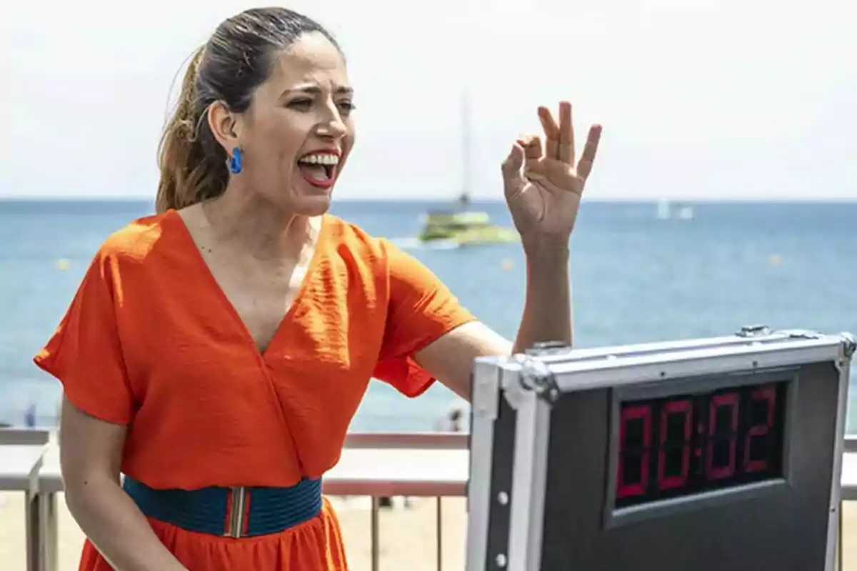 Fotografía de Nuria Marín con un vestido naranja en Tens un minut? gesticulando con la mano frente a un cronómetro que marca 00:02, con el mar y un barco de fondo.