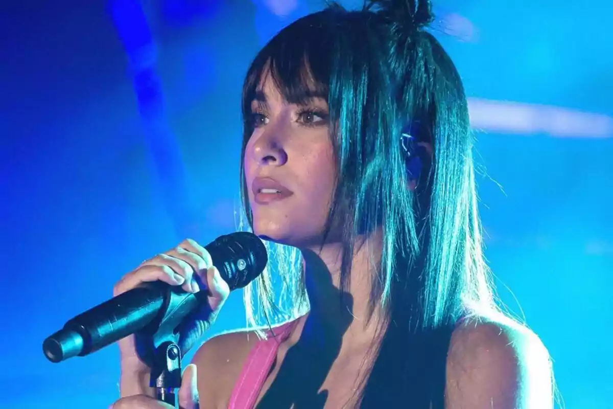 Una dona amb cabell llarg i fosc sosté un micròfon mentre canta en un escenari il·luminat amb llums blaves. Una dona amb cabell llarg i fosc sosté un micròfon mentre canta en un escenari il·luminat amb llums blaves.