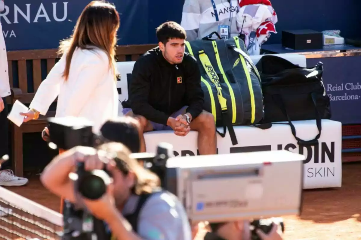 Un hombre está sentado en un banco junto a una cancha de tenis, rodeado de bolsas deportivas, mientras una mujer y varios fotógrafos están cerca.