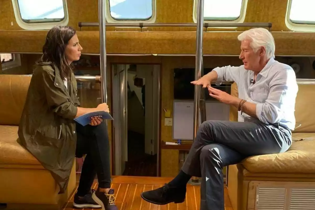 Ana Pastor i Richard Gere conversen asseguts en un sofà dins d'un vaixell, la dona sosté un quadern mentre l'home gesticula amb les mans a El Objetivo.