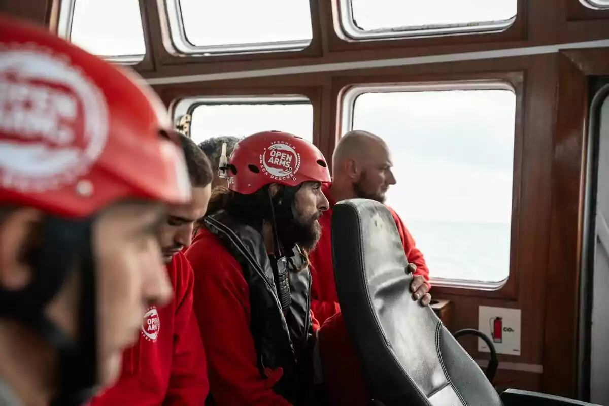 Personas con cascos rojos y ropa roja observan desde el interior de un barco a través de las ventanas.