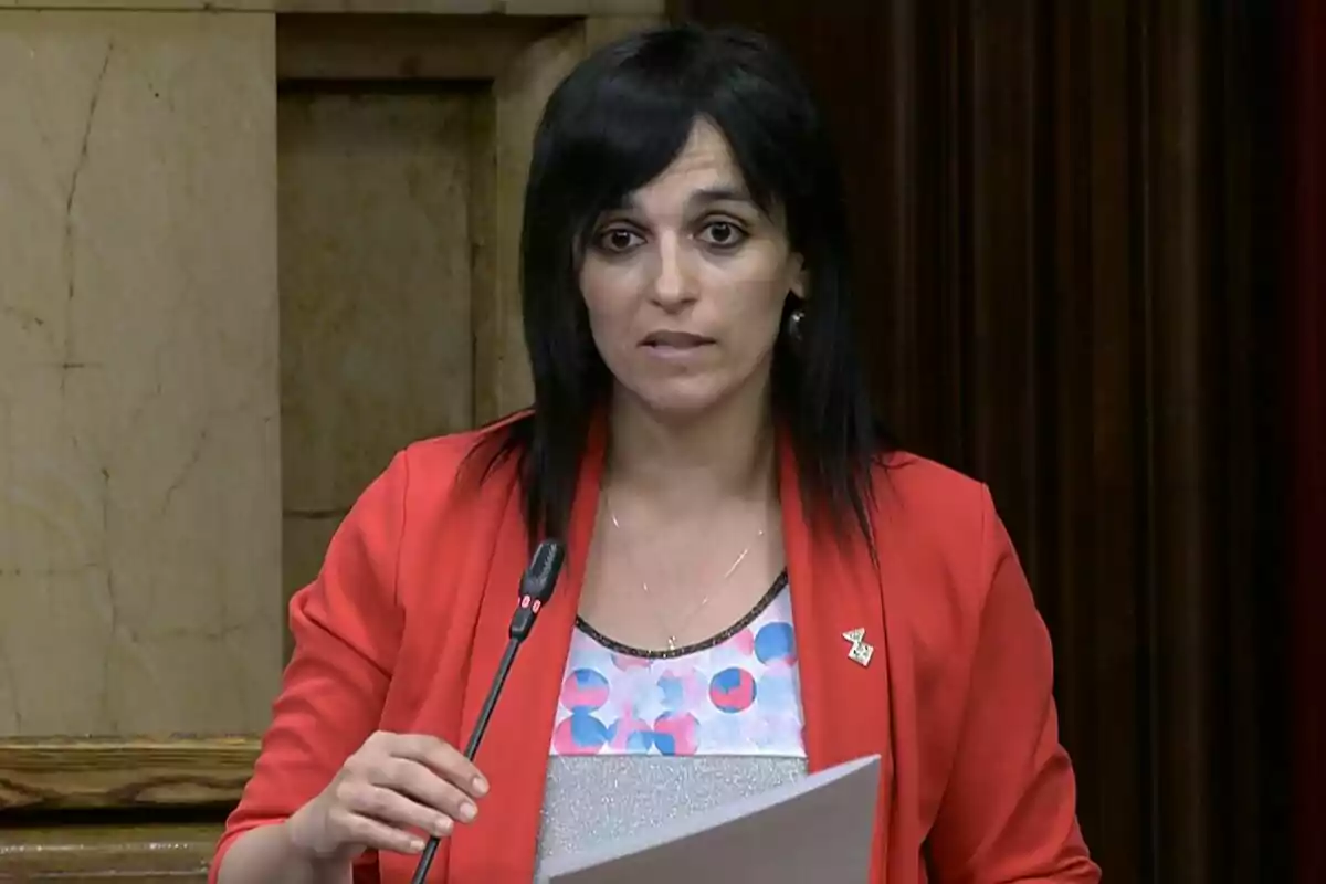 Dark-haired woman with straight hair speaking into a microphone while holding some papers, dressed in a red jacket and patterned blouse, in an institutional setting.