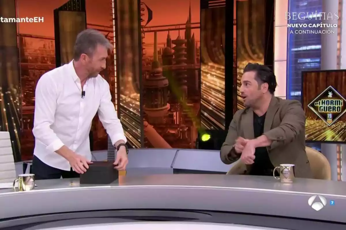 Pablo Motos i David Bustamante conversen animadament a El Hormiguero amb un fons futurista i una pantalla que mostra el logotip d'un programa.