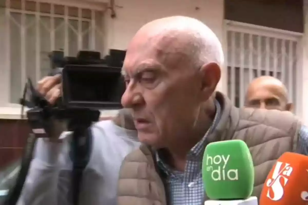 Un home gran amb jaqueta encoixinada és entrevistat per diversos mitjans de comunicació, amb micròfons de colors davant seu i una càmera de vídeo al fons. Un home gran amb jaqueta encoixinada és entrevistat per diversos mitjans de comunicació, amb micròfons de colors davant seu i una càmera de vídeo al fons.