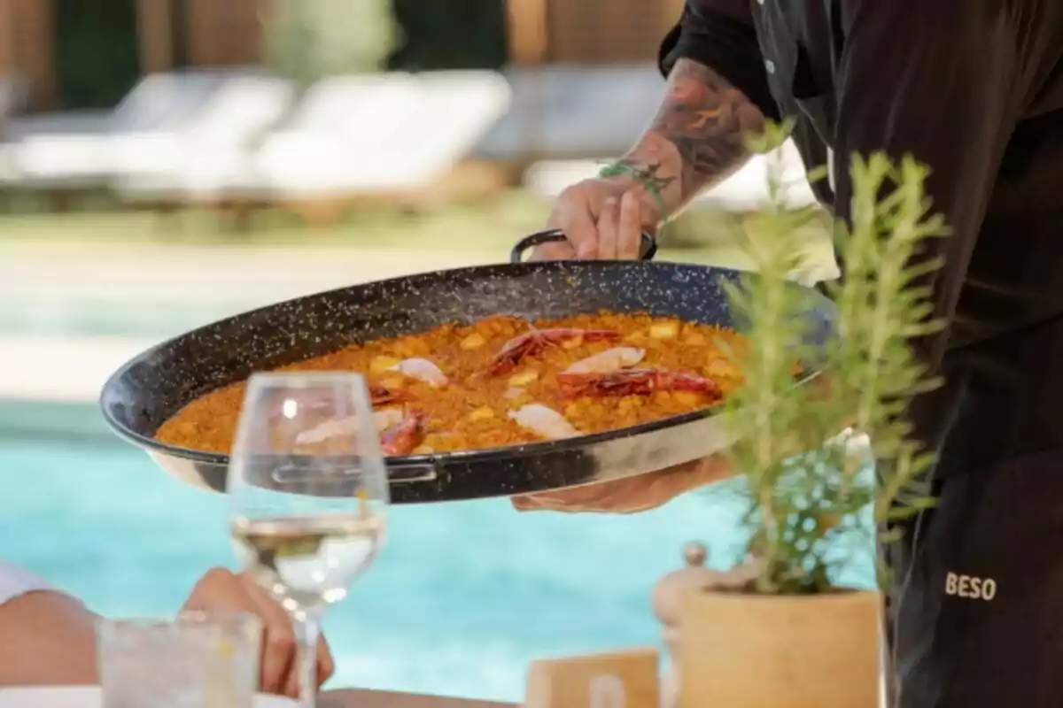 A waiter serving paella by a swimming pool, with a glass of wine in the foreground.