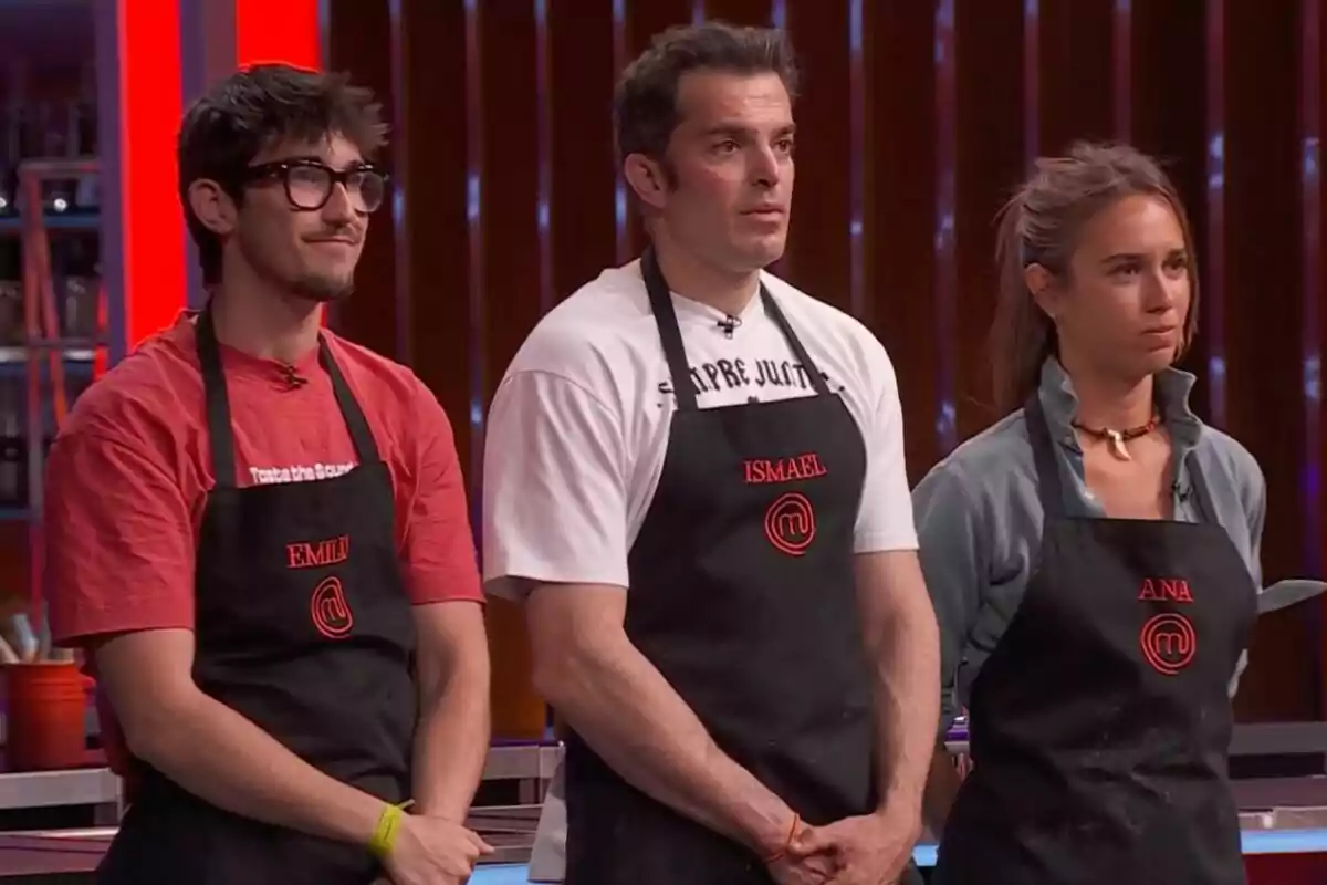 Tres concursants de 'Masterchef' estan drets un al costat de l'altre portant davantals negres amb els seus noms brodats en vermell i el logotip del programa, tots miren cap al davant amb expressions serioses.