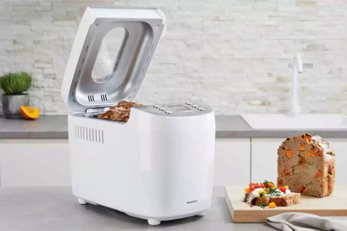 Bread maker with the lid open in a modern kitchen, next to a cutting board with freshly baked bread and slices decorated with vegetables. Bread maker with the lid open in a modern kitchen, next to a cutting board with freshly baked bread and slices decorated with vegetables.