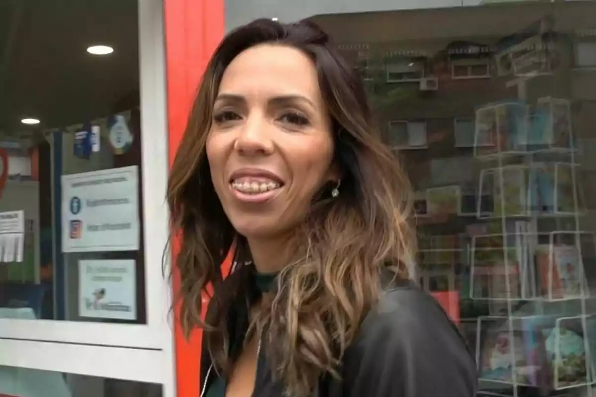 Paola Olmedo sonriente con cabello castaño claro y ondulado está de pie frente a una tienda con una puerta de vidrio y un estante de revistas en el fondo. Paola Olmedo sonriente con cabello castaño claro y ondulado está de pie frente a una tienda con una puerta de vidrio y un estante de revistas en el fondo.
