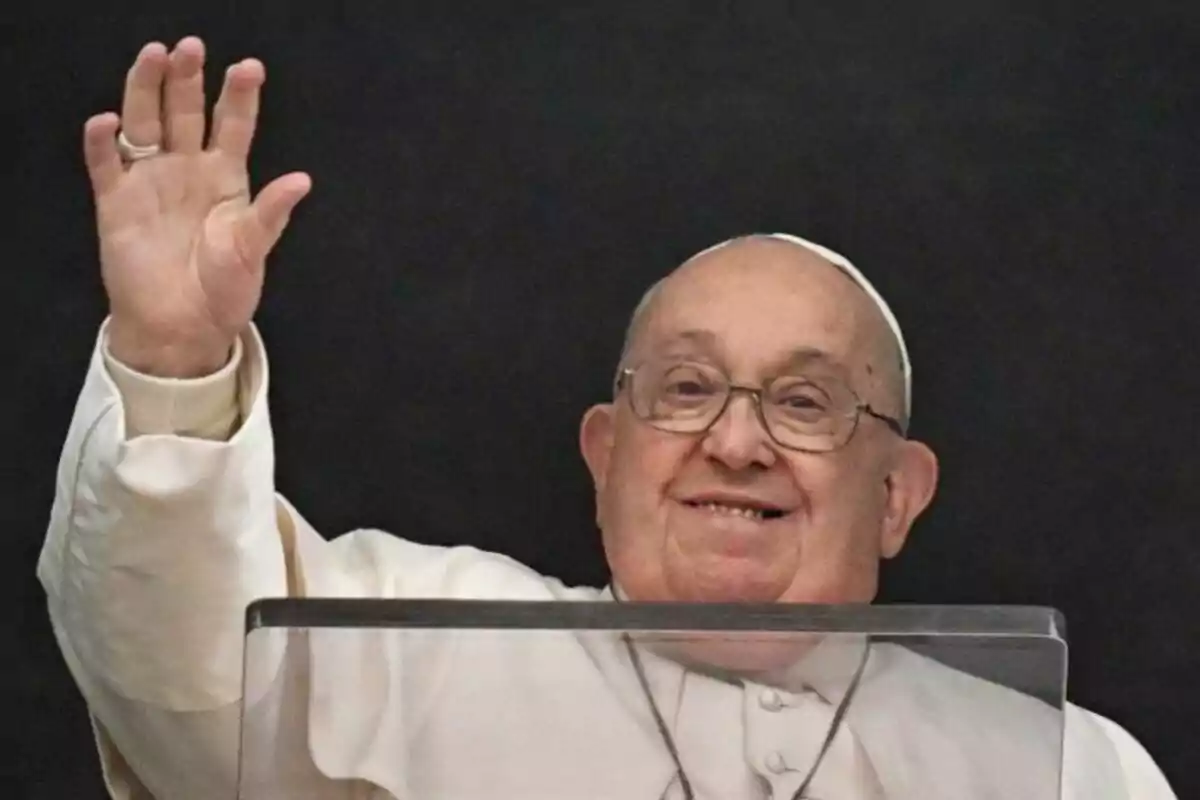 Plano corto del Papa Francisco con gafas y vestimenta blanca levantando la mano y sonriendo detrás de un atril.