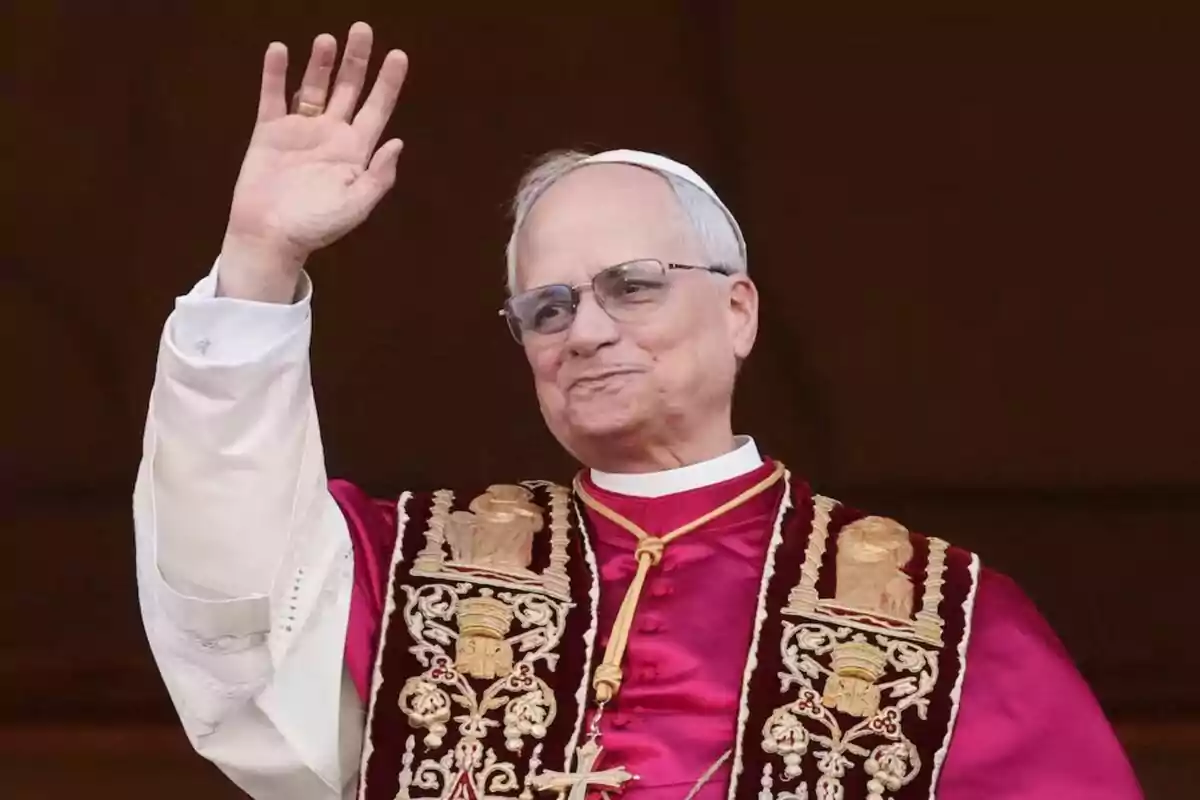An elderly man with glasses and religious attire raises his hand in greeting.