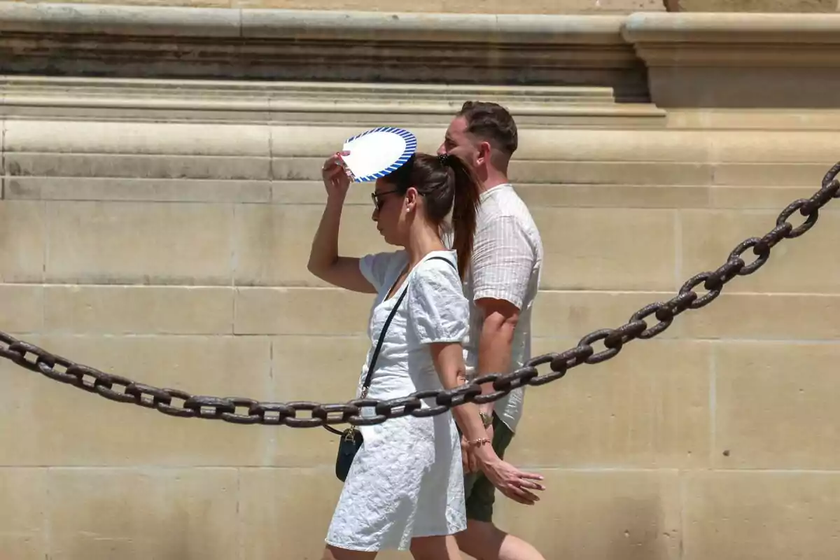Dues persones caminen al costat d’una paret de pedra mentre una d’elles es protegeix del sol amb un ventall. Dues persones caminen al costat d’una paret de pedra mentre una d’elles es protegeix del sol amb un ventall.