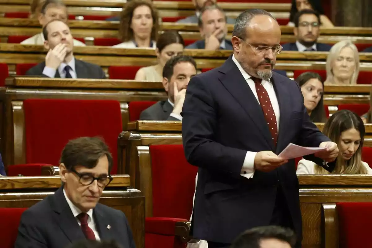 Alejandro Fernández dret amb vestit i corbata vermella sosté un document mentre salvador Illa està assegut al seu escó al Parlament de Catalunya