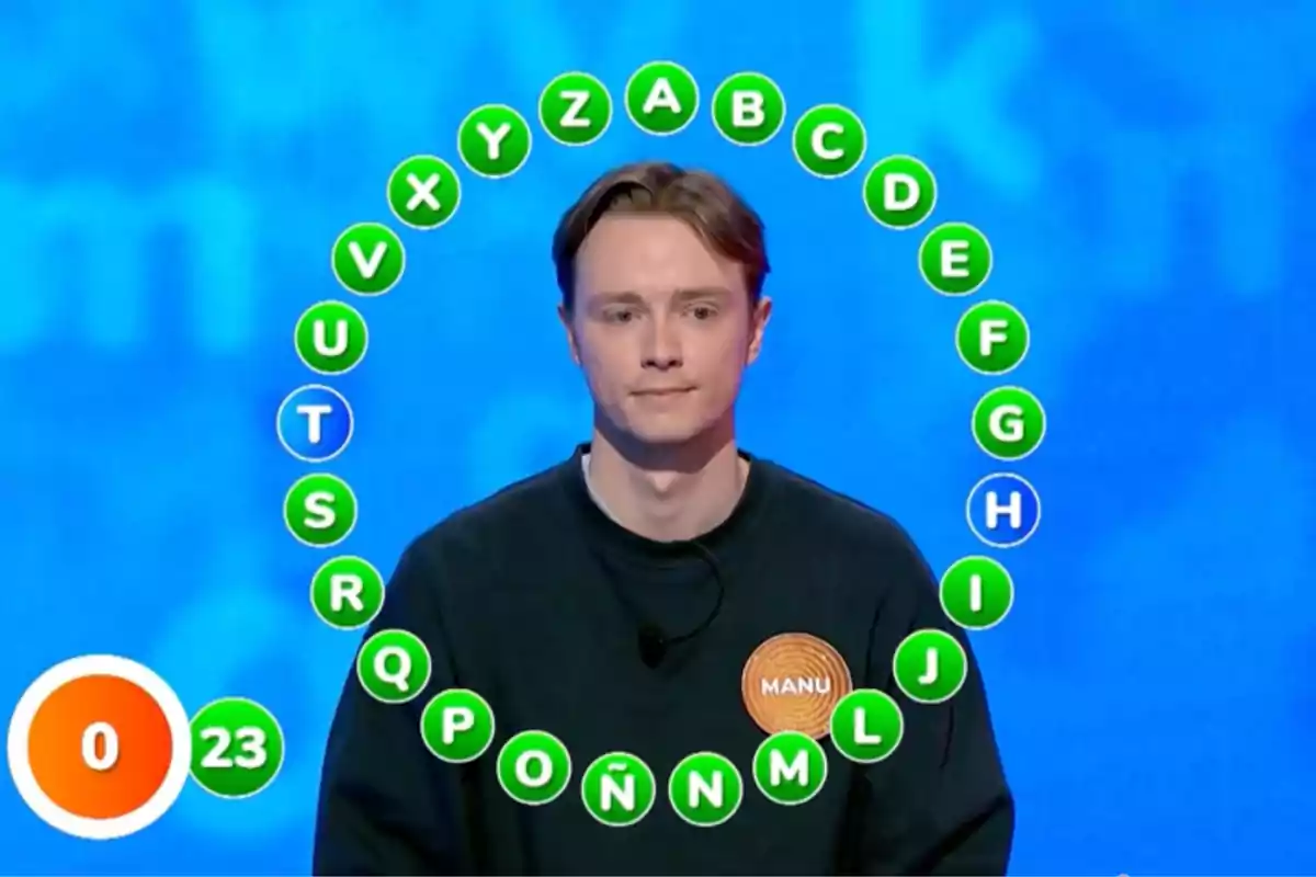 Manu is standing in front of a blue background with a circle of green letters and some white ones around him, wearing a dark jersey with a label that says Manu, and in the bottom left corner there is a scoreboard showing the number 0 and a timer at 23 seconds. Screenshot from 'Paspalabra' Manu is standing in front of a blue background with a circle of green letters and some white ones around him, wearing a dark jersey with a label that says Manu, and in the bottom left corner there is a scoreboard showing the number 0 and a timer at 23 seconds. Screenshot from 'Paspalabra'
