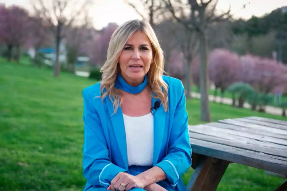 Patricia Cerezo asseguda en un banc de fusta en un parc amb arbres i gespa, vestint un vestit blau i brusa blanca a Aquí es fa. Patricia Cerezo asseguda en un banc de fusta en un parc amb arbres i gespa, vestint un vestit blau i brusa blanca a Aquí es fa.