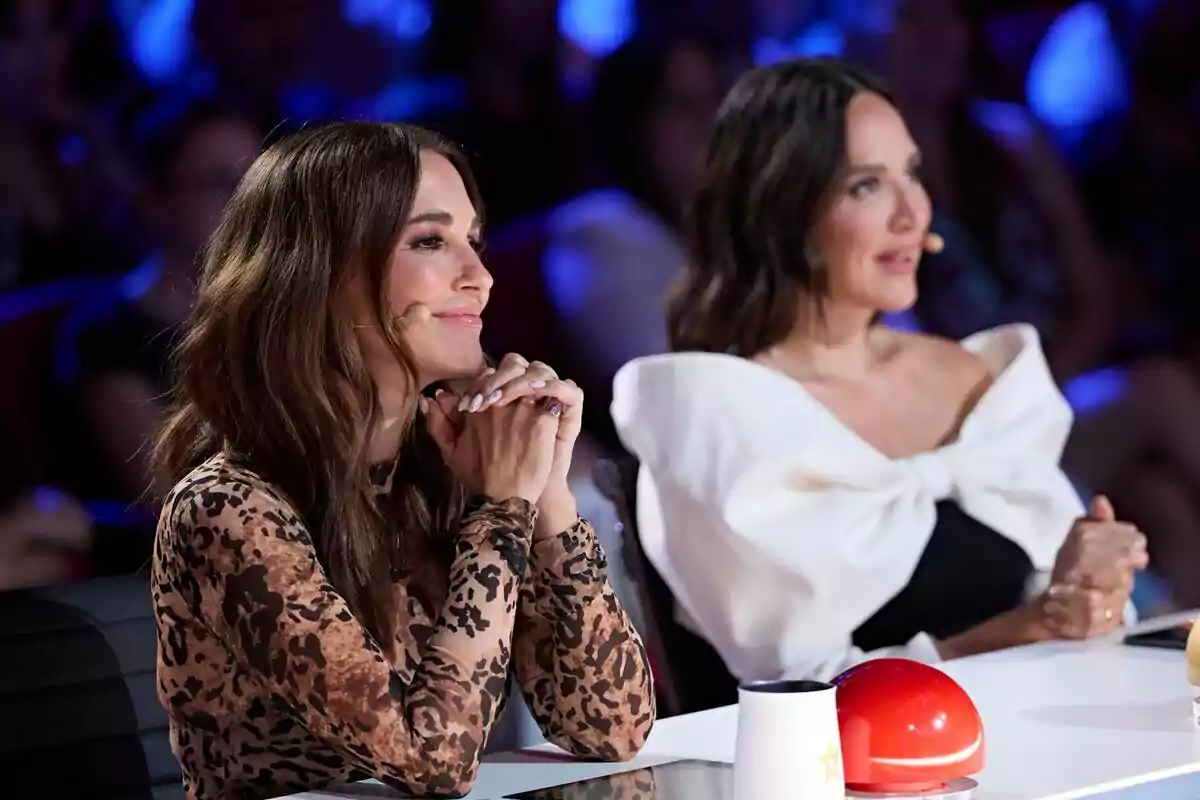 Paula Echevarría amb Tamara Falcó assegudes a la taula de Got Talent, una amb un vestit d'estampat animal i l'altra amb un vestit blanc amb un gran llaç, totes dues amb micròfons a l'orella i una llum blava de fons.