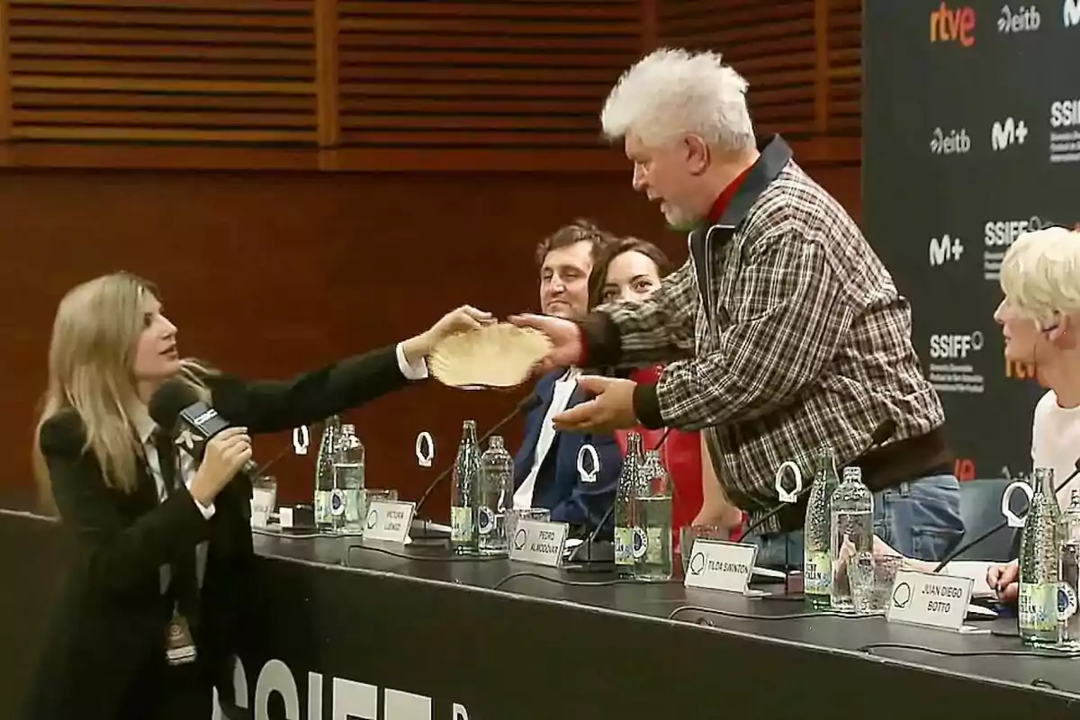 Paula Púa com a reportera de Caiga quien caiga lliura un objecte a Pedro Almodóvar durant una roda de premsa, mentre altres persones observen des de la taula.