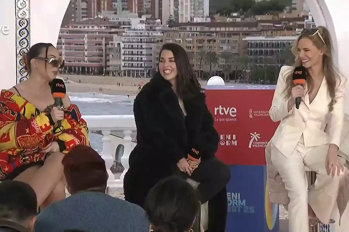 Paula Vázquez, Ruth Lorenzo i Inés Hernand sostenen micròfons de RTVE mentre conversen en la presentació del Benidorm Fest a l'aire lliure amb un fons de platja i edificis.