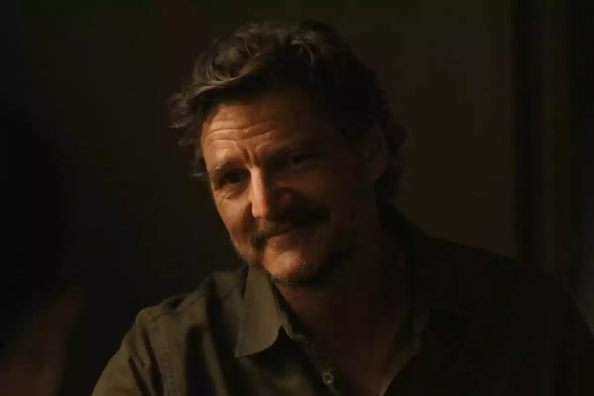 Pedro Pascal con barba y cabello rizado sonríe suavemente en un ambiente con poca luz en la segunda temporada de The Last of us.