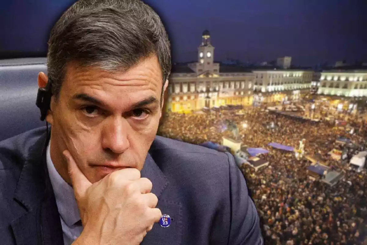 Pla mig curt de Pedro Sánchez amb cara de preocupació i amb la mà dreta a la barbeta i de fons una imatge aèria de l'acampada del 15M a la Porta del Sol de Madrid de nit