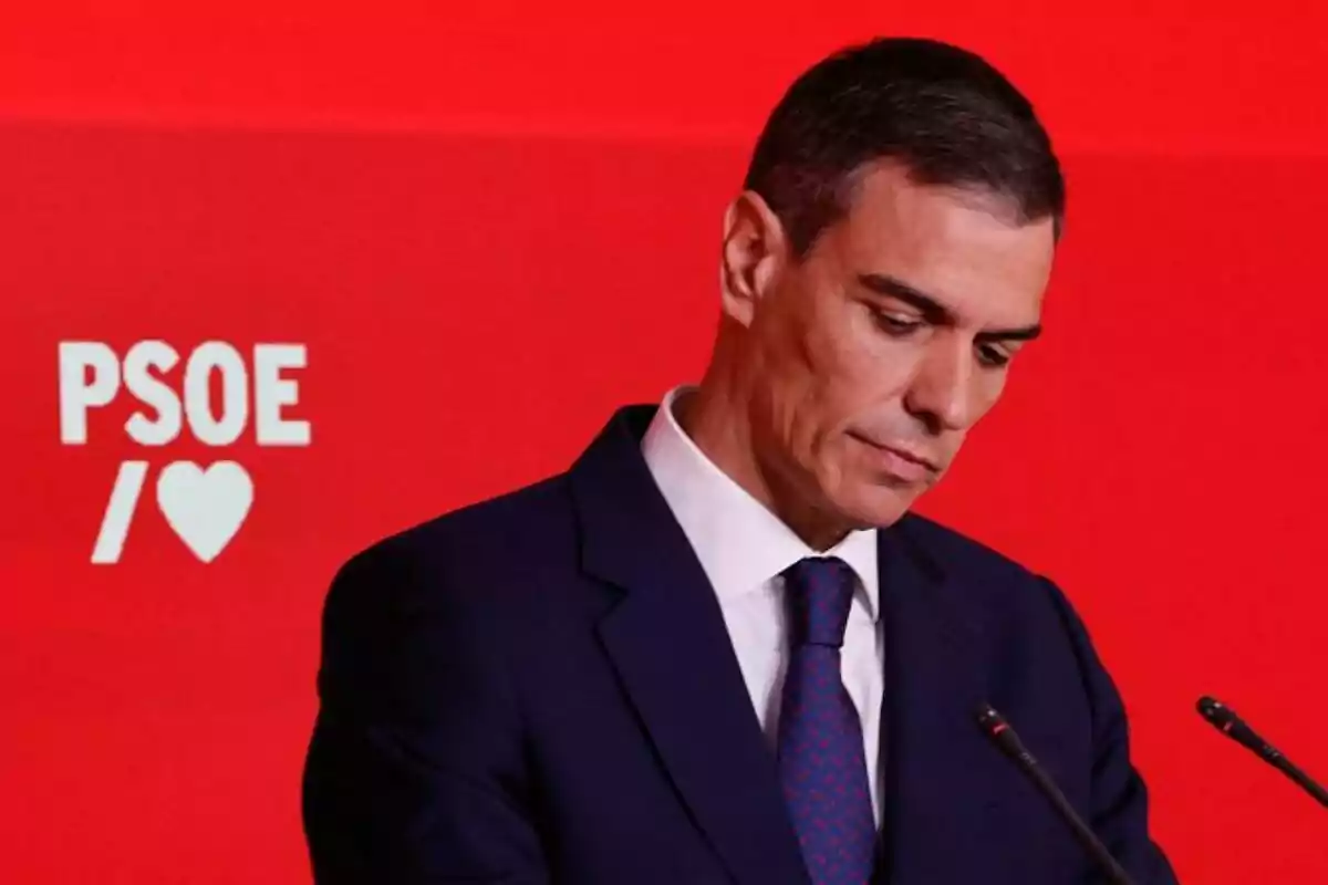 Home amb vestit fosc i corbata blava davant d’un fons vermell amb el logotip del PSOE i un cor blanc Home amb vestit fosc i corbata blava davant d’un fons vermell amb el logotip del PSOE i un cor blanc