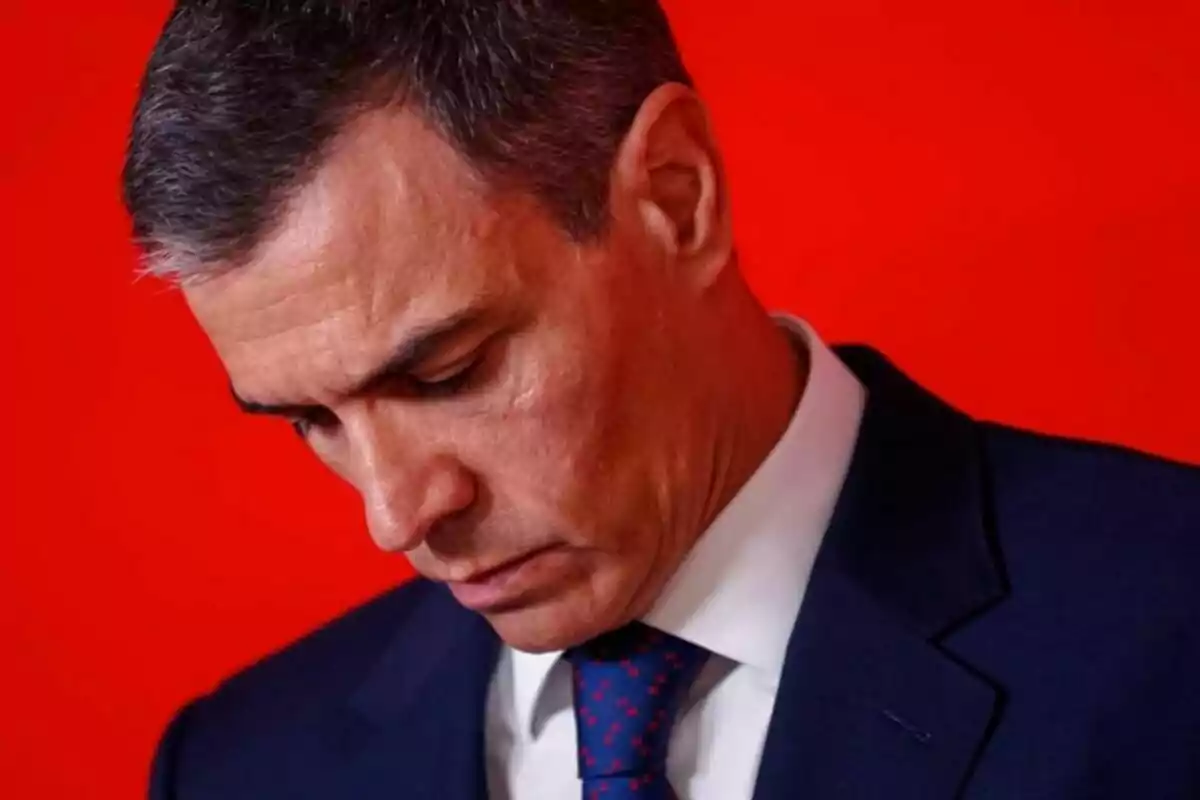 Short-haired, gray-haired man in a blue suit and tie looking down against a red background