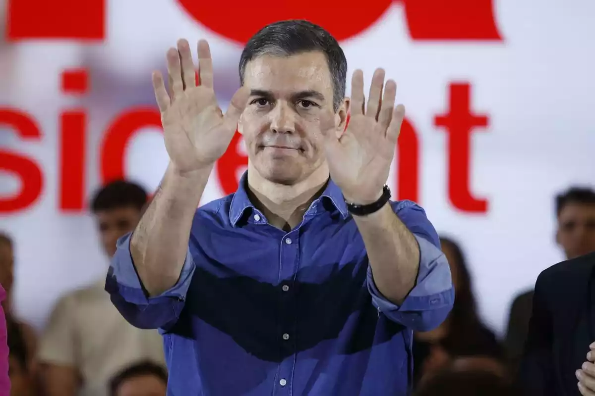 Pla mitjà de Pedro Sánchez en un míting a Catalunya, amb camisa blava i amb els dos braços i mans cap endavant saludant